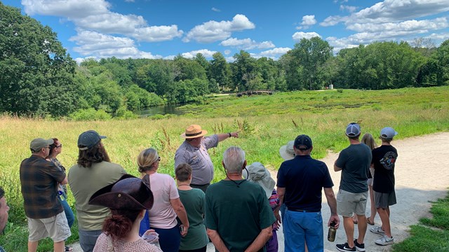 A park ranger in flat hat stands in front of a visitor group. The ranger points at a wide landscape.