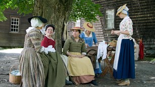 Five women in colonial dress sit under a tree near a wooden house. The women are spinning thread.