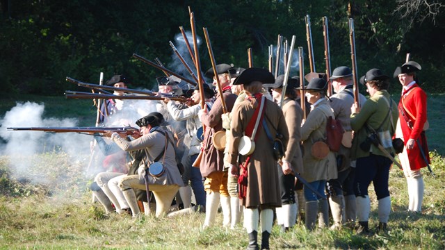 Group of 40 men dressed as Revolutionary War militia soldiers firing their muskets, white smoke.