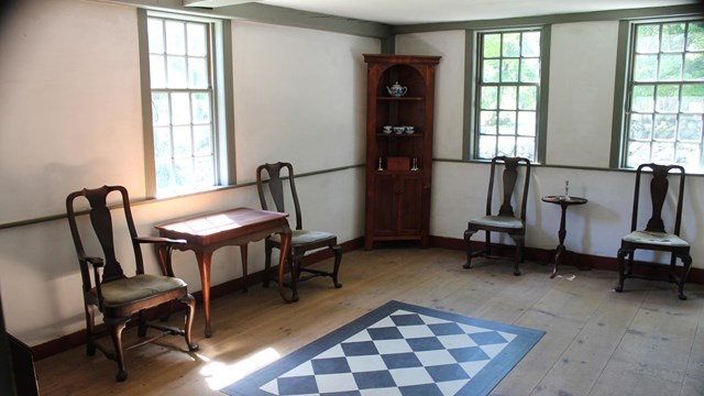 A room in a historic house with a wooden floor, checkerboard floor cloth, side chairs and a table