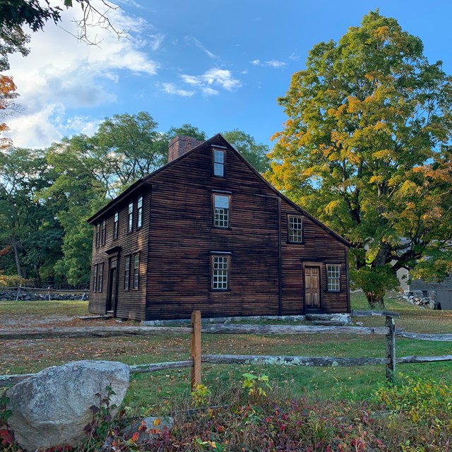 A wooden colonial house sits surrounded by trees and stonewalls