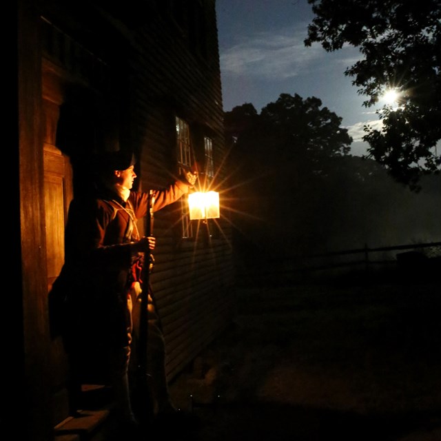 An armed colonial solider stands in the doorway of a wooden house at night holding a lantern