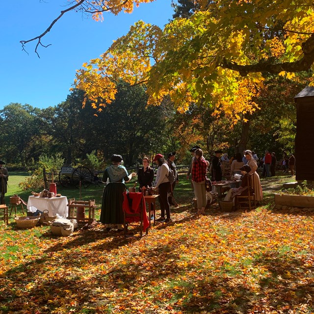 A group of visitors talk with 18th century living historians under trees turning red and orange