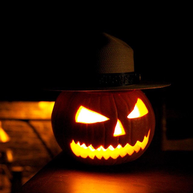 A Jack-'o'-Lantern with Ranger flat hat on sits on a table before a colonial fireplace 