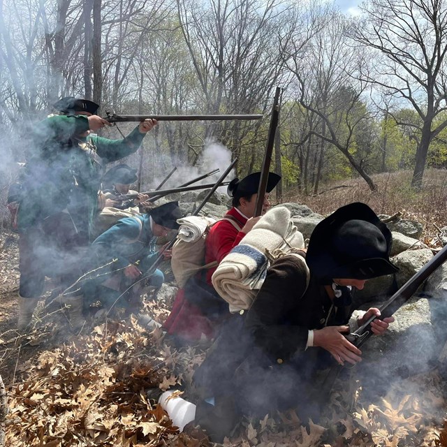Militia soldiers crouch behind a stone wall loading and firing muskets while surrounded by smoke.