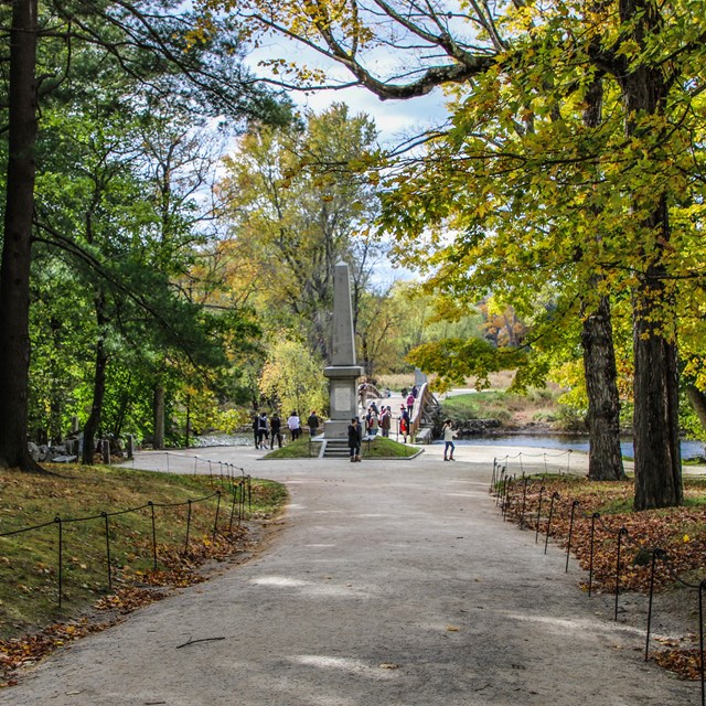 A paved walkway extends between rows of trees to an obelisk shaped monument.