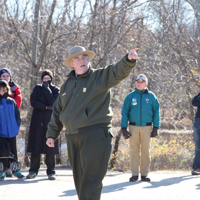 A National Park Ranger surrounded by visitors points off camera while speaking