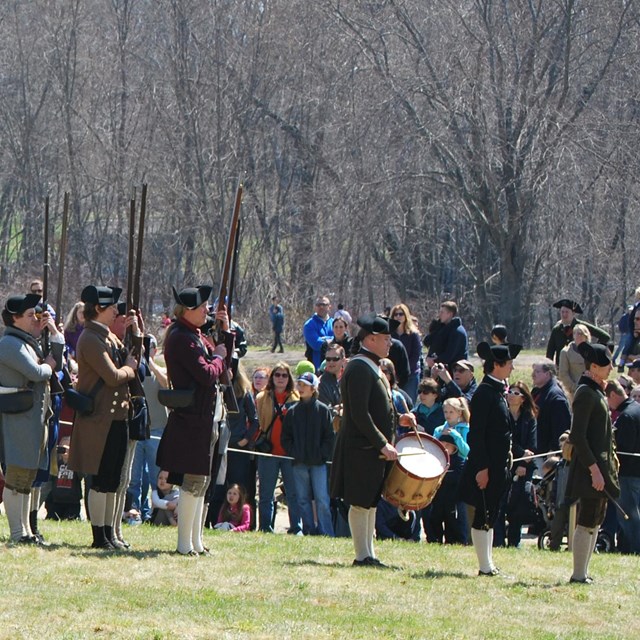 Colonial Militia stand in an open field and practice drilling with muskets in front of a large crown