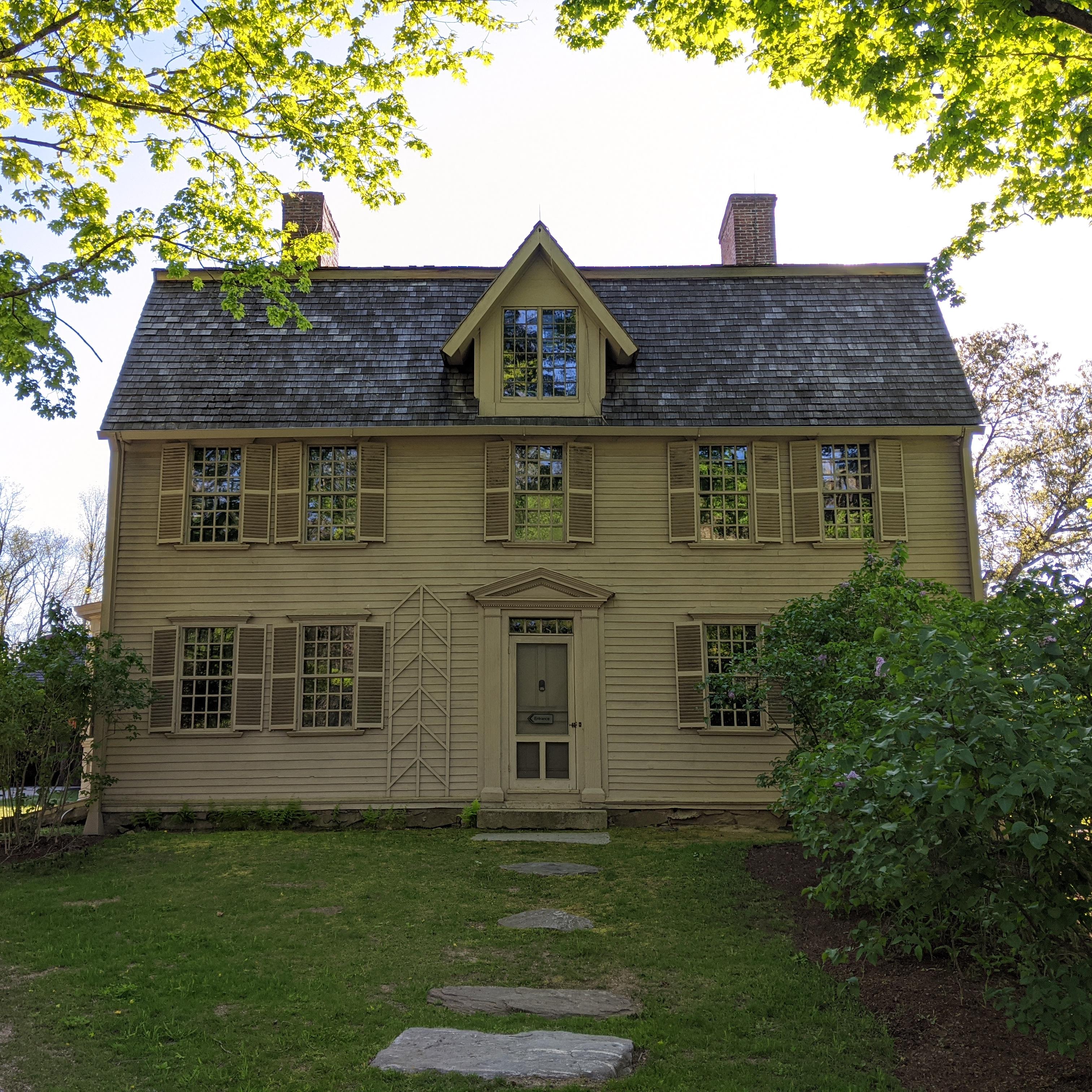 A two and a half story wooden colonial house with a center doorway. Trees flank the house.