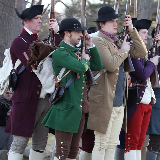 colonial reenactors, dressed in non-uniform clothing, perform drill with their muskets