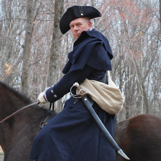 Man in colonial clothing, black cocked hat, dark blue coat and a sword sits on a brown horse.