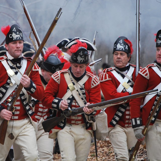 A group of red coated British soldiers advance forward with muskets at the ready