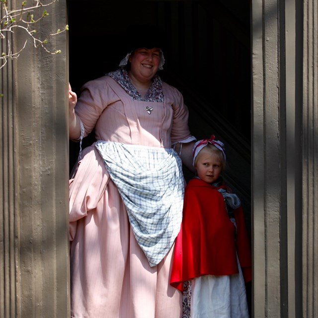woman and young girl dressed in colonial clothing stand in the doorway of a brown colonial house