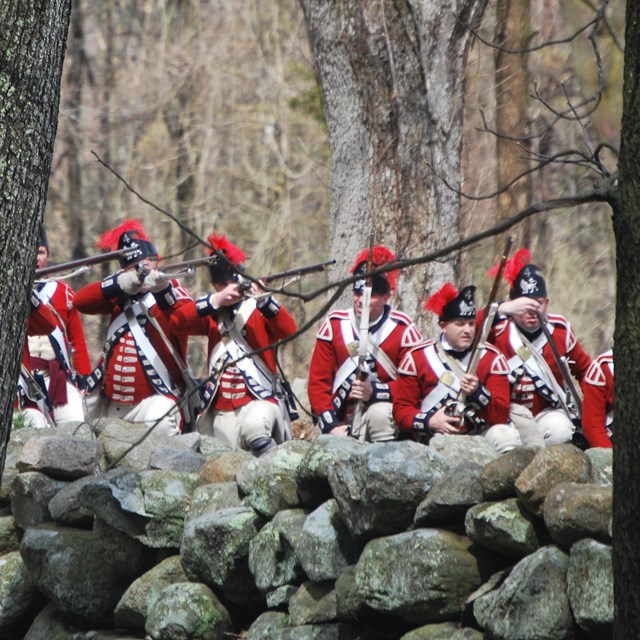Red-coated British soldiers of the American Revolution prepare to fire from behind a stone wall