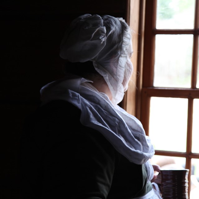 A woman in colonial dress with a white cap and neckerchief stares out a multi-paned window.