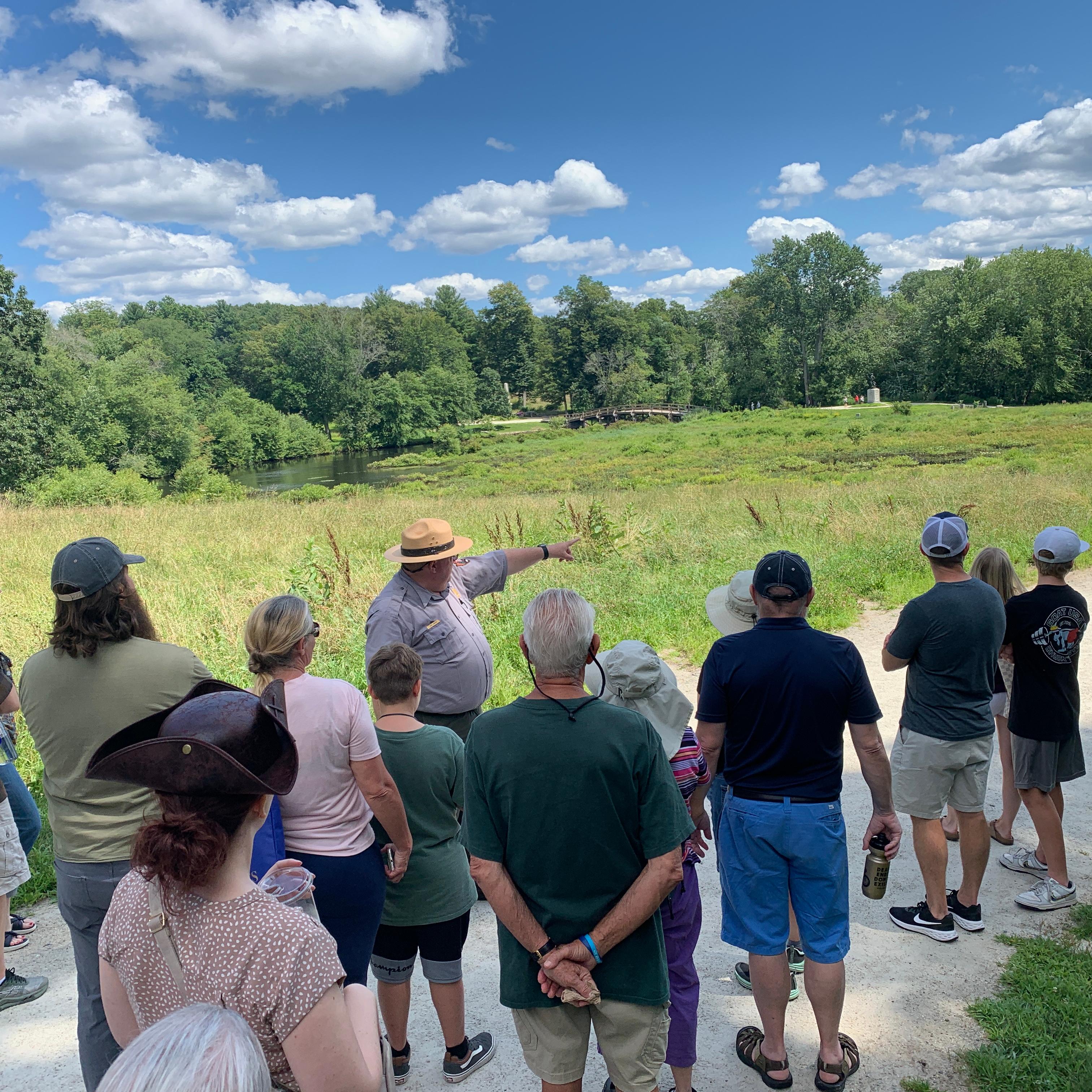 A park ranger in flat hat stands in front of a visitor group. The ranger points at a wide landscape.