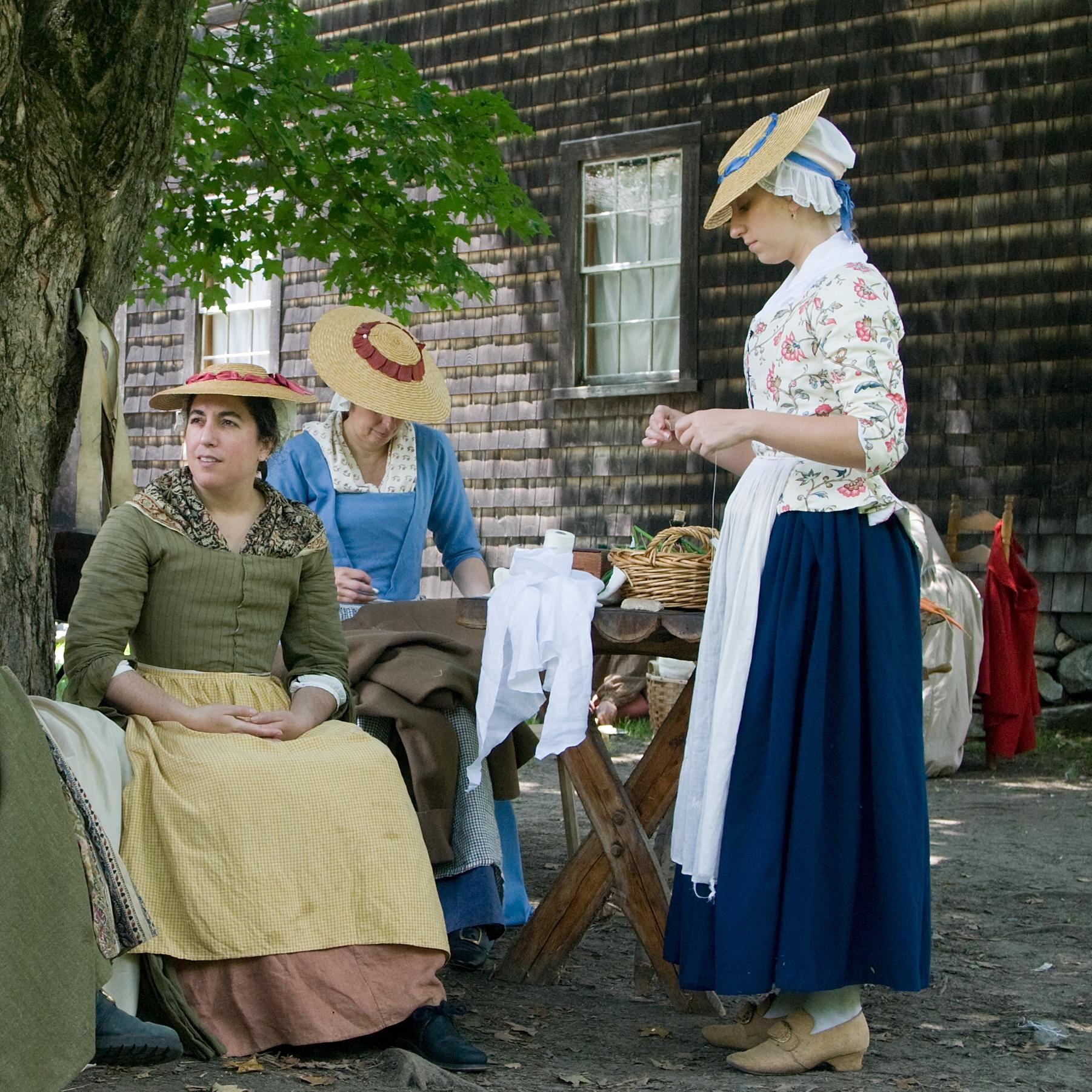 Five women in colonial dress sit under a tree near a wooden house. The women are spinning thread.