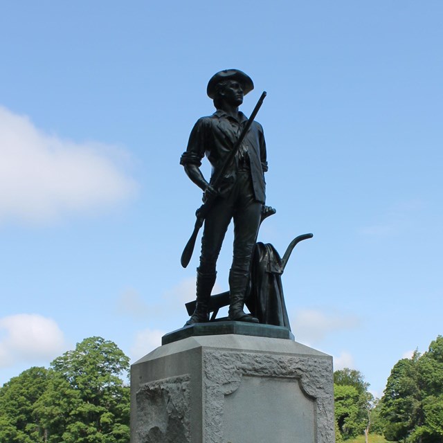 A bronze statue of a minute man sits atop a granite base. The man is walking and carrying a musket