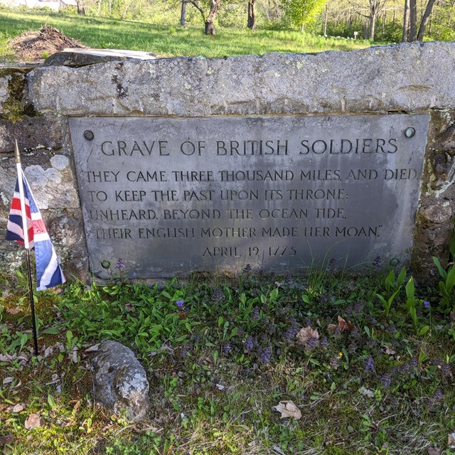 A gravestone is imbedded into a low stonewall surrounded by fencing and green grass.