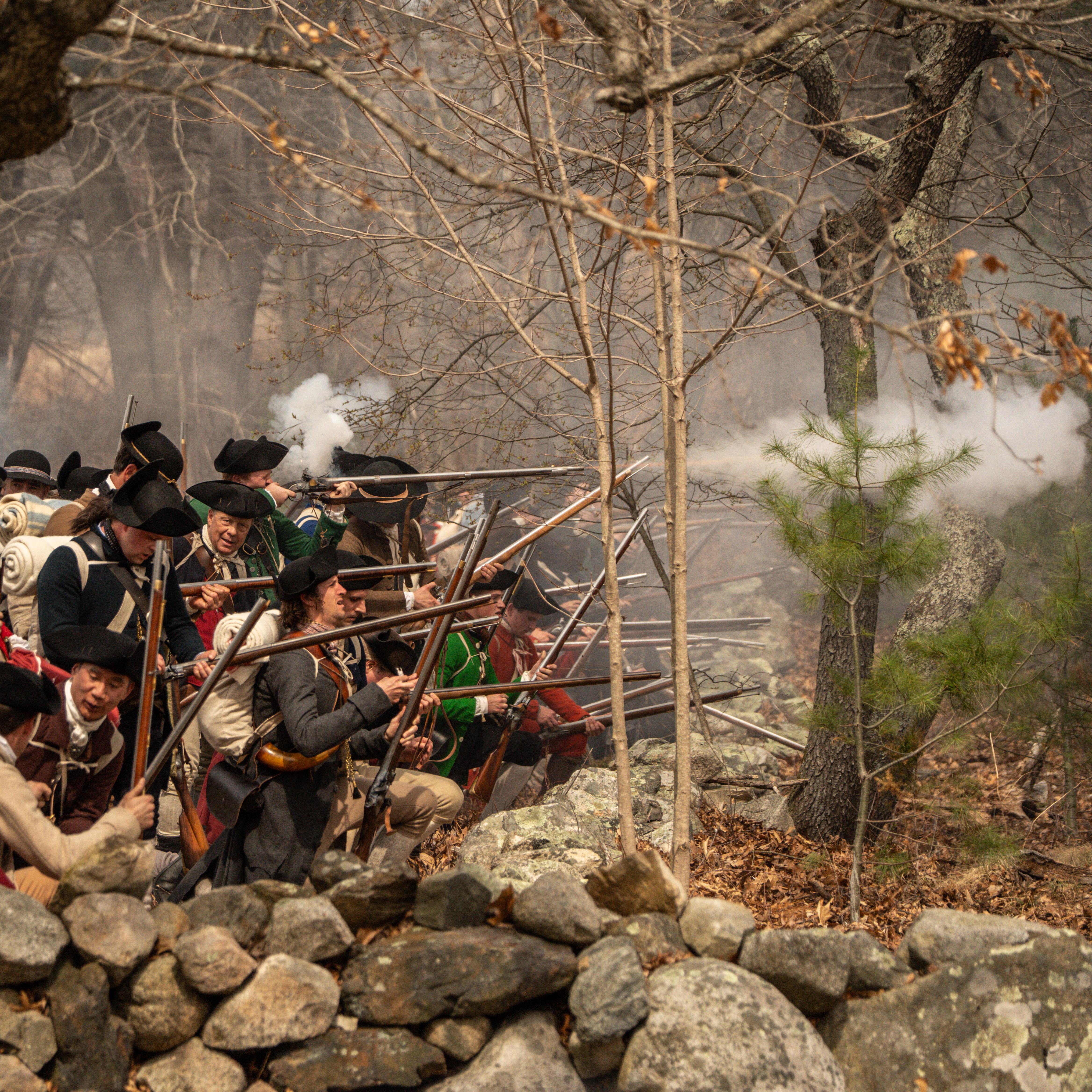 Militia soldiers crouch behind a stone wall loading and firing muskets while surrounded by smoke.
