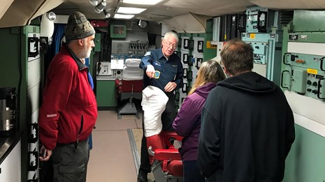 A group of people in a small room with electronic panels.