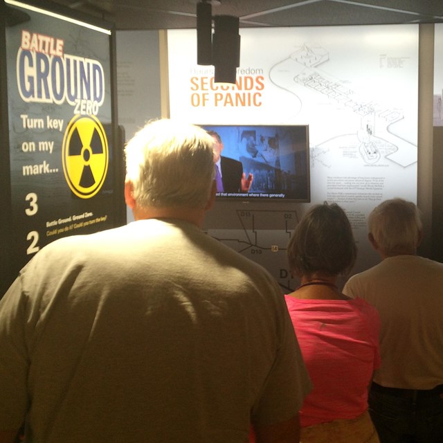 Visitors in the exhibit area watch a program on a screen. 