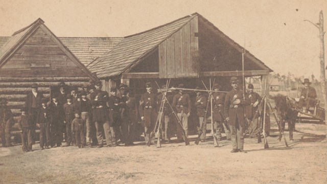 A sepia-toned image of soldiers with stacked muskets in front of log building.