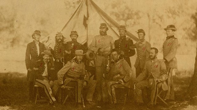 A sepia-toned image of officers standing and sitting in front of tent.