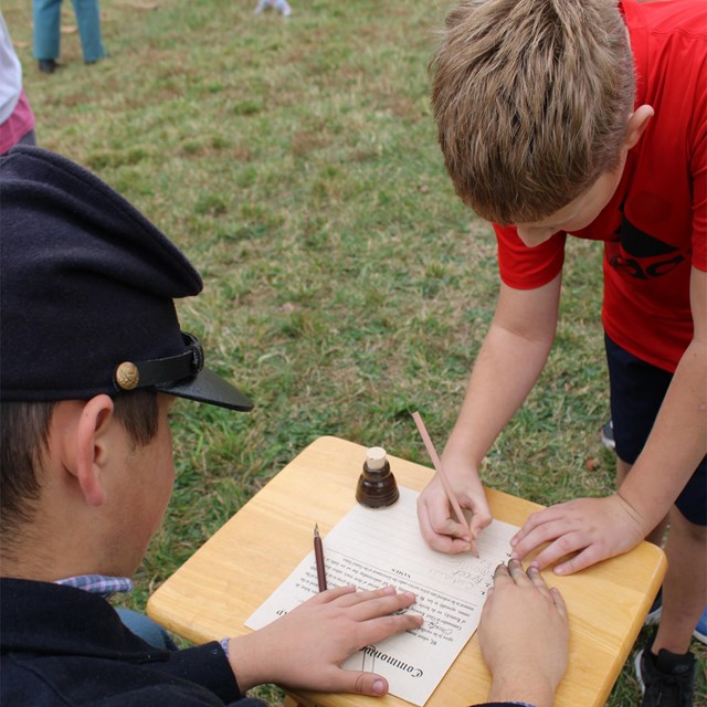 A male youth leaning down signing a fake Civil War enlistment paper.