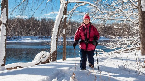 A hiker in a red coat and hat hikes through a snow-covered forest with a river in the background.