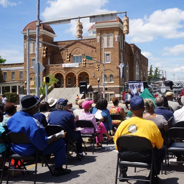 Crowd in chairs on a street in front of a brick church