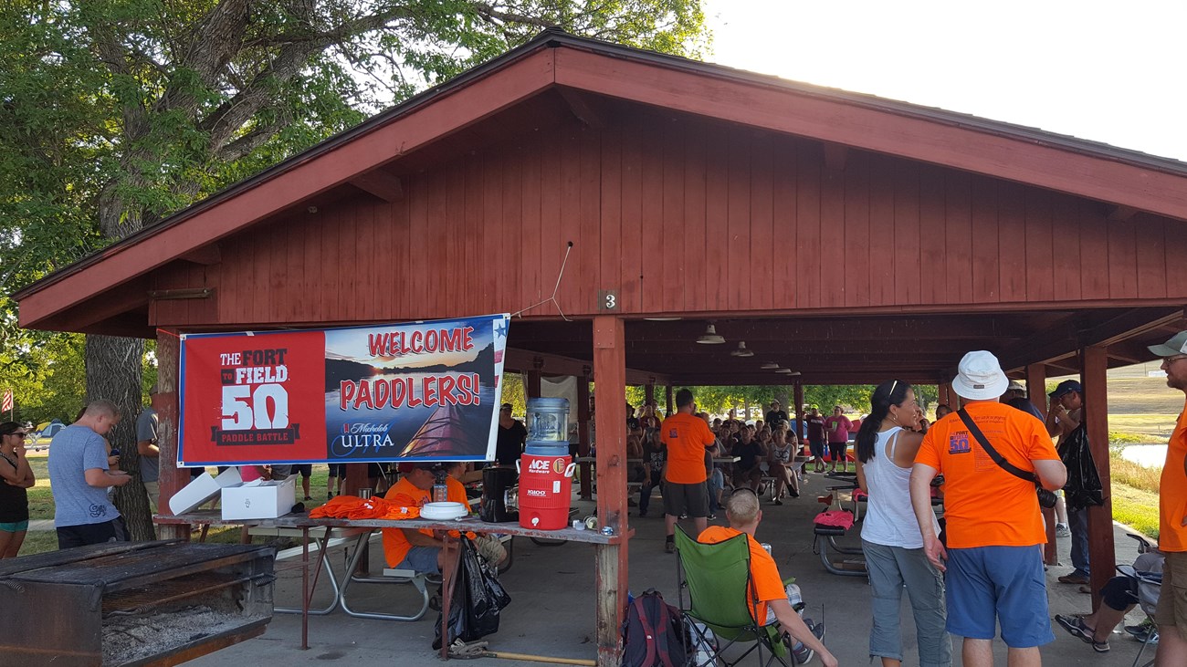 About 50 people gather under a large red picnic shelter waiting for an event to start.