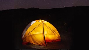 Yellow tent illuminated by the light inside. Mountains in the background with stars in the sky.
