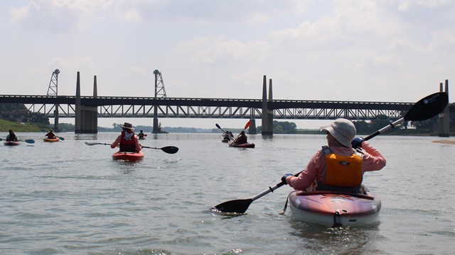 A view from a kayak of other paddlers kayaking towards a bridge spanning a large calm river