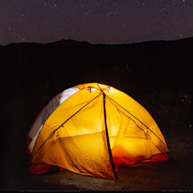 Yellow tent illuminated by the light inside. Mountains in the background with stars in the sky.