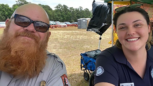 Woman in a white polo sitting next to a man with a red beard sitting under a tent.