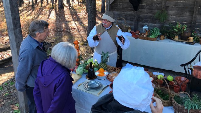 Park Volunteer in Living History Clothing with food on a table talking with three visitors.