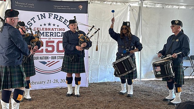 Five people in plaid kilts and blue over shirts standing under a white tent