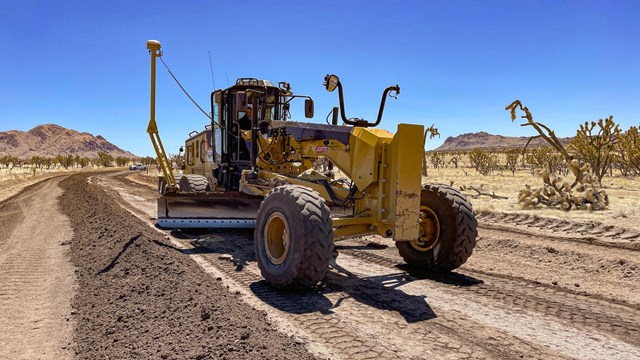 A yellow road grader with two GPS antennas on the blade moves earth material along a road