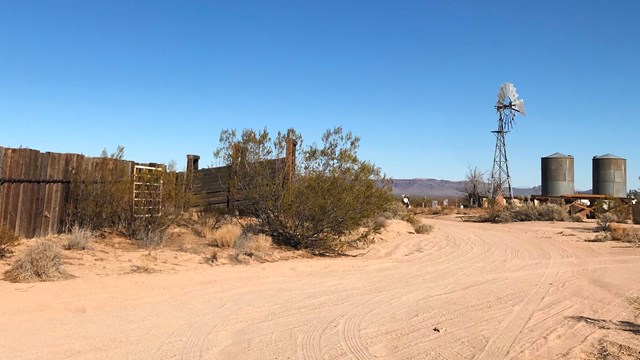 A dirt road with a wooden corral, windmill, and metal tanks.