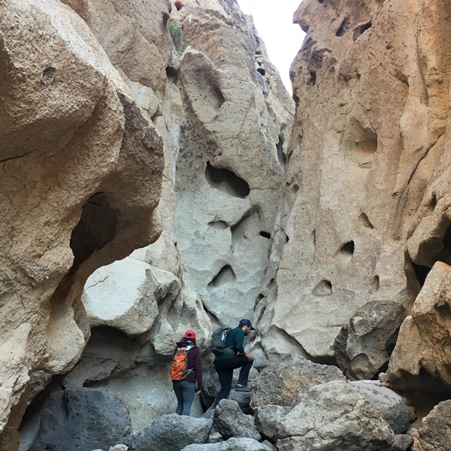 Two hikers on the Rings Tails in Banshee Canyon. Canyon walls are tall and filled with many holes.