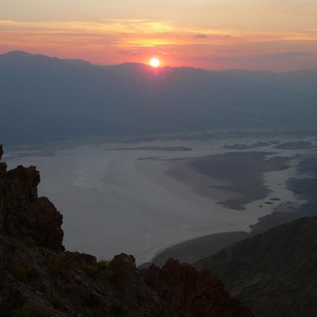 Sunset over the Death Valley Basin with mountains, clouds, and a sandy desert floor below.
