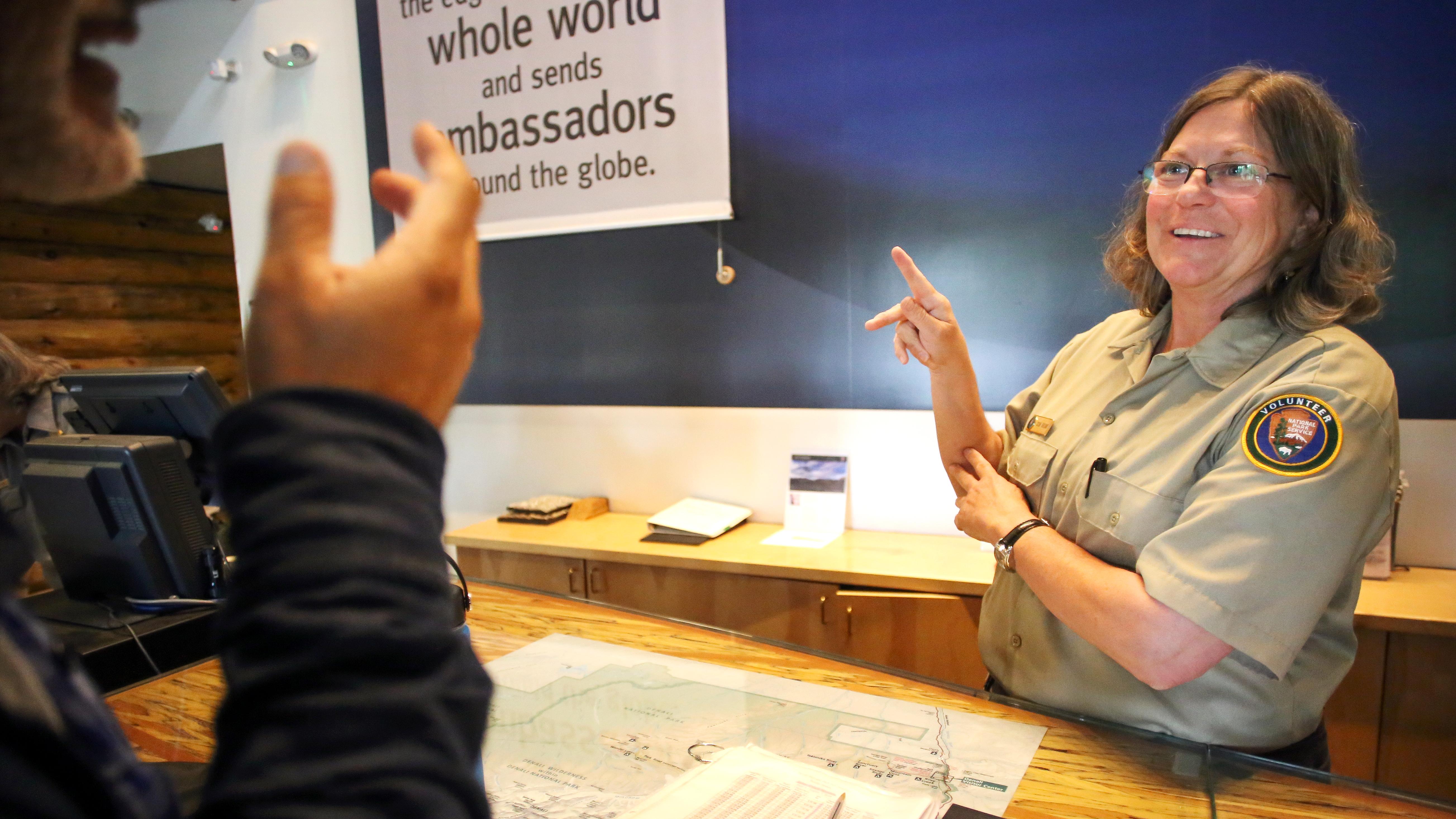 A park volunteer behind a desk communicates using sign language with a visitor.