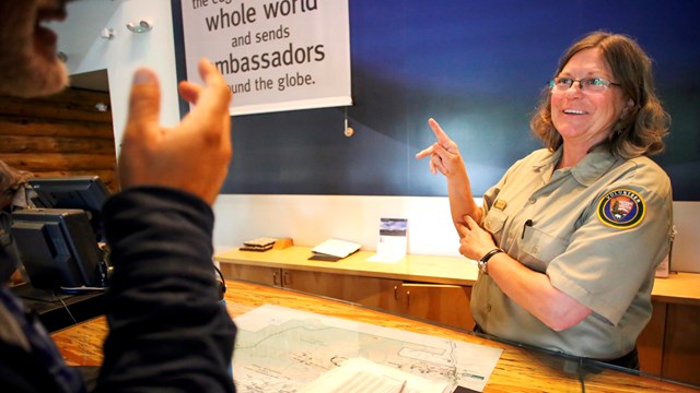 A park volunteer behind a desk communicates using sign language with a visitor.