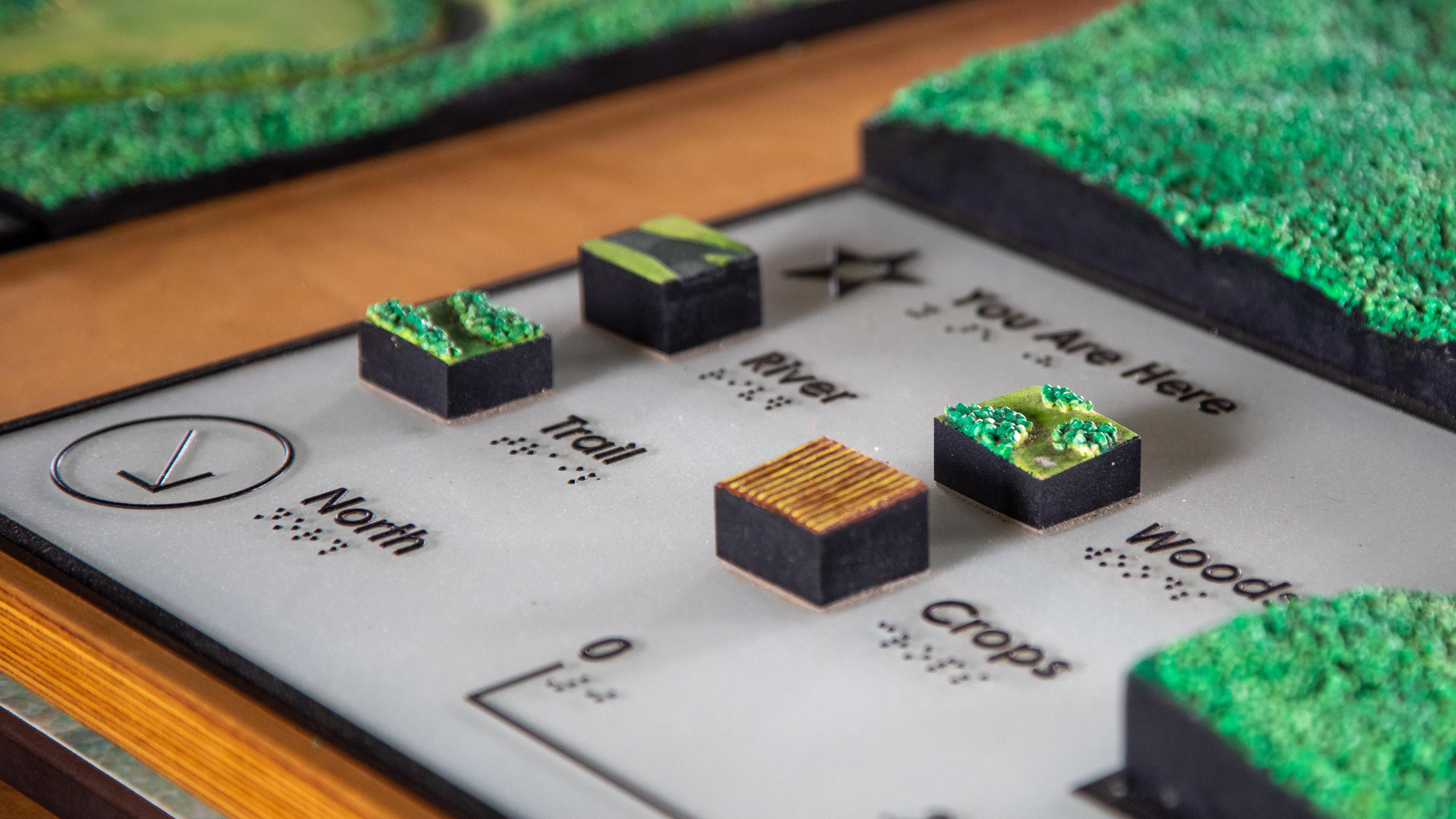 A tactile map key showing textures for river, trail, crops, and woods with braille labels.