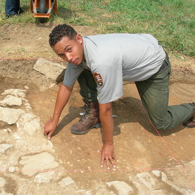 Park archeologist kneels in a large square hole. He points to a feature made of stones.