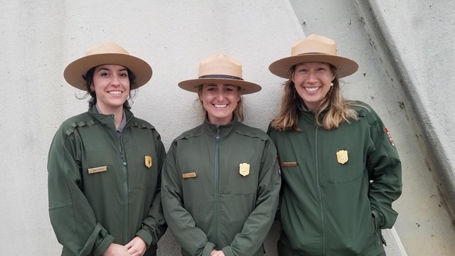 Three uniformed park rangers stand shoulder to shoulder. 