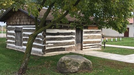 A small log cabin, with white chinking, with a green grass lawn.