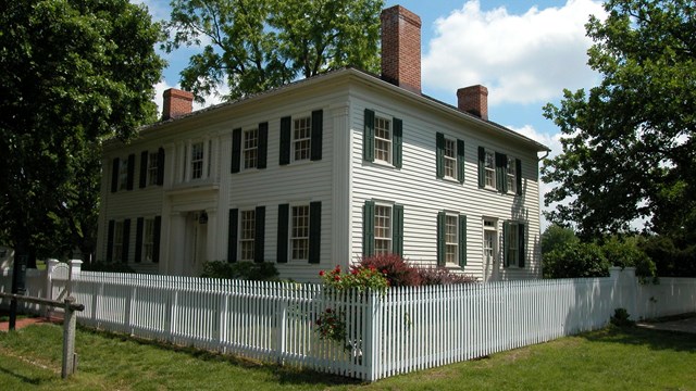 A large, two-story, white traditional house, with a white picket fence and a green lawn.