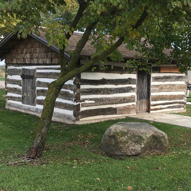 A small log cabin, with white chinking, with a green grass lawn.